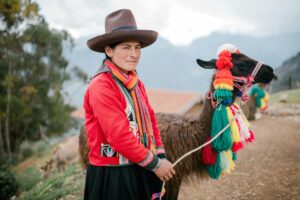 A woman in traditional attire stands with a colorfully decorated llama in the Peruvian Andes.